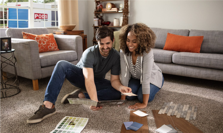 A happy couple is sitting on the floor of their living room, looking at paint, flooring, and tile samples for their home renovations. A PODS portable storage container is visible through the window.