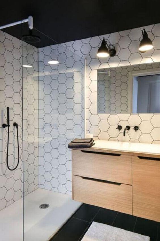 Banheiro com azulejo hexagonal branco por todo banheiro, piso branco na area do box e piso preto fora, armário amadeirado com pia branca, torneira e chuveiro preto e espelho retangular