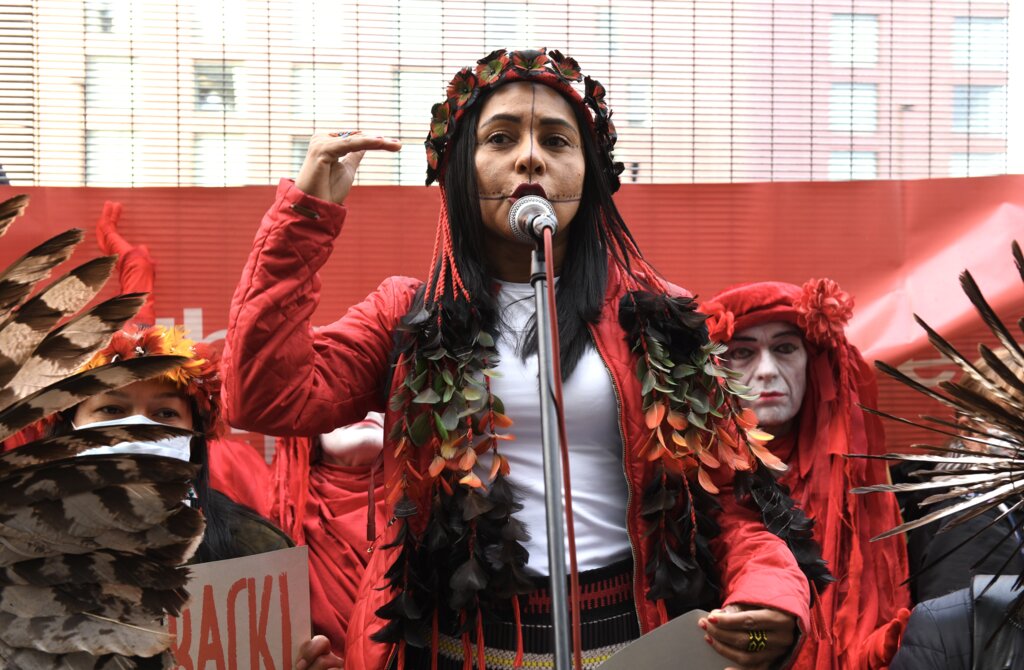 A young indigenous woman speaks to a rally. She wears a feather garland and jacket and has lines drawn down her face.