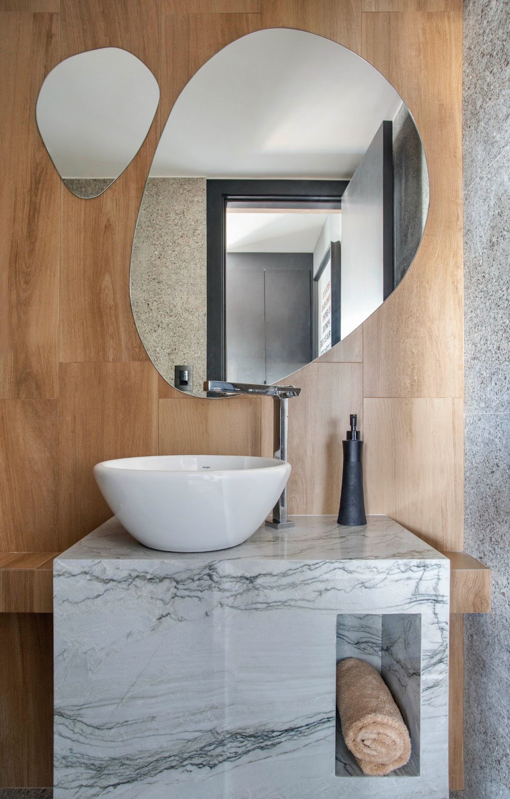Banheiro com espelho em formato livre aplicado sobre a parede de porcelanato com padrão de madeira, bancada de mármore com cuba branca.