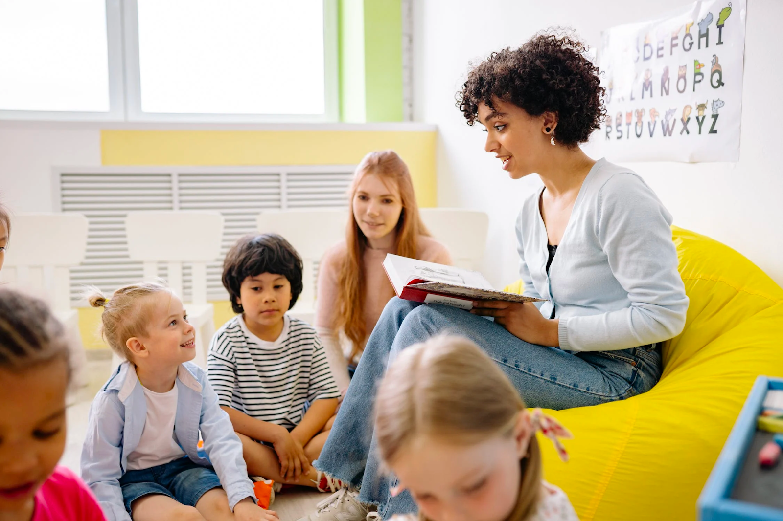 preschool teacher sitting on a yellow bean bag reading to a group children seated on the floor