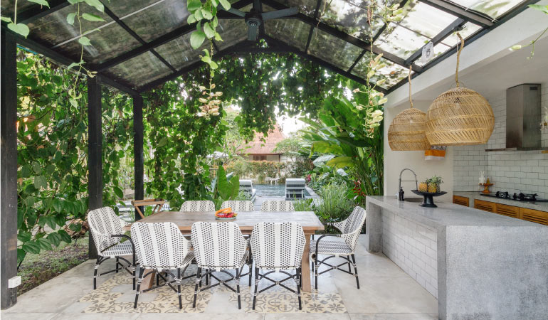 A modern patio with a full outdoor kitchen and a covered dining space. Lush green vines hang all around the patio, creating privacy. 