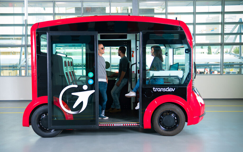 An electric shuttle, round and red, sits three people on it. They're smiling. There is no driver because the vehicle is self-driving. It is in an airport, where people need transporting efficiently, especially those with additional needs.