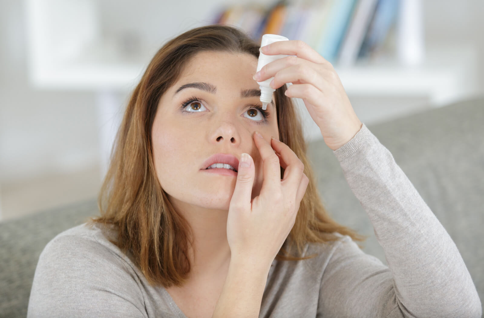 A woman holding a small bottle of eye drops in her left hand and putting them on her left eye.