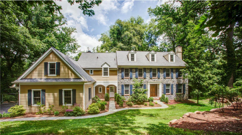 A yellow siding and stone, two-story home in Newtown Square, Pennsylvania. The lawn is well manicured and there are trees casting shade on the house from three sides. 