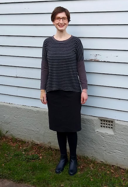 Siobhan stands in front of a weatherboard house. She wears a drop shoulder knit tee with grey/black striped body and concrete grey neckband and sleeves, with black straight knee length skirt, black leggings and ankle boots. She is smiling.