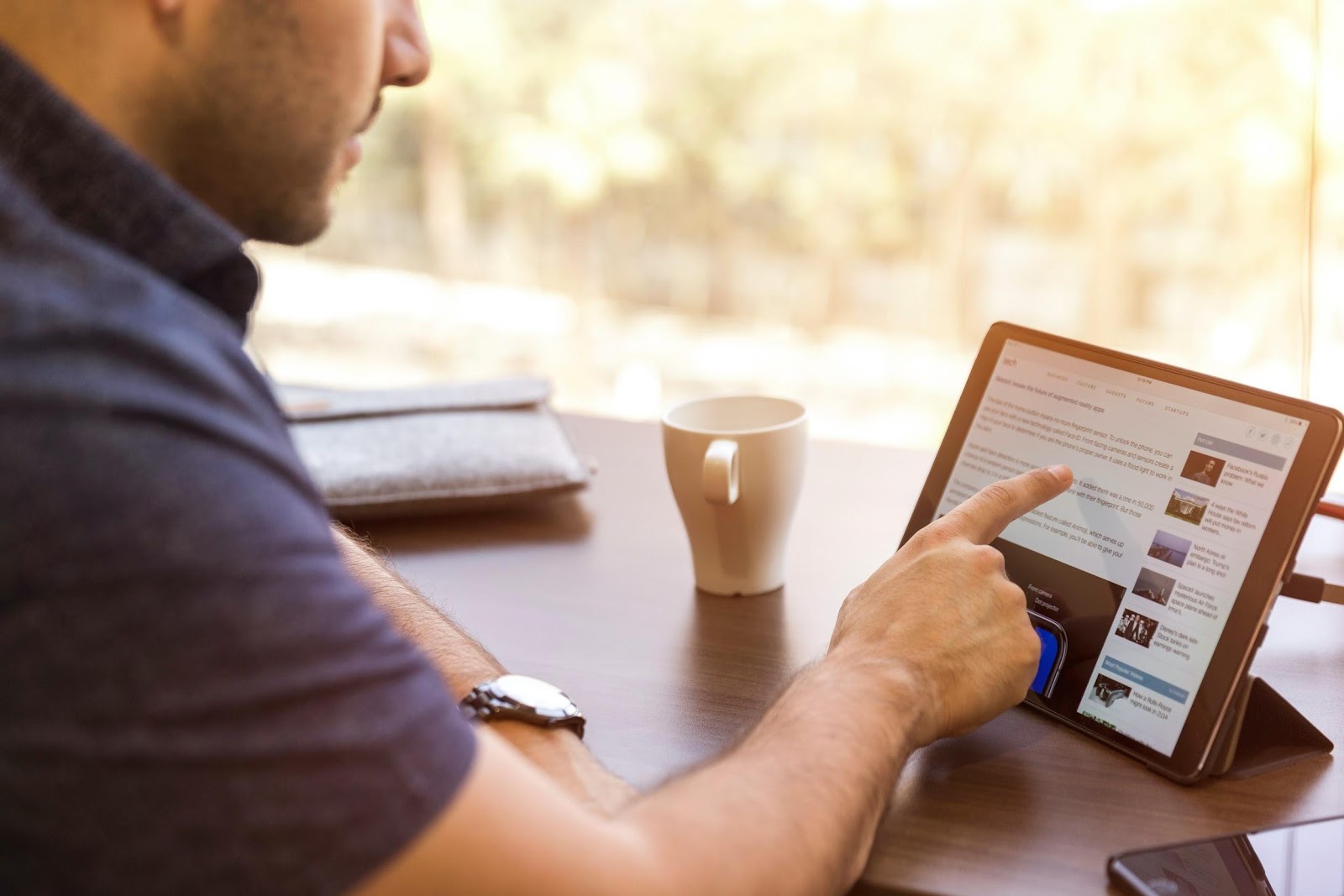 Hombre usando un tablet, que está encima de una mesa de madera. Junto al tablet, hay una taza de café, un teléfono celular y una carpeta.