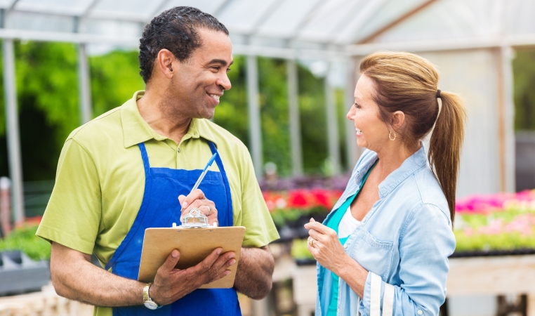 A woman is smiling as she speaks with an associate at a local plant nursery. The associate is also smiling and is wearing a blue apron and holding a clipboard. 