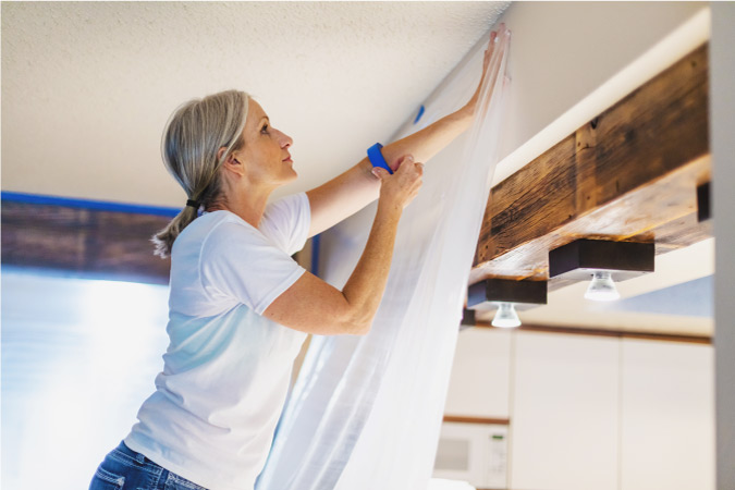 A woman taping plastic to a wall while preparing to paint her living room