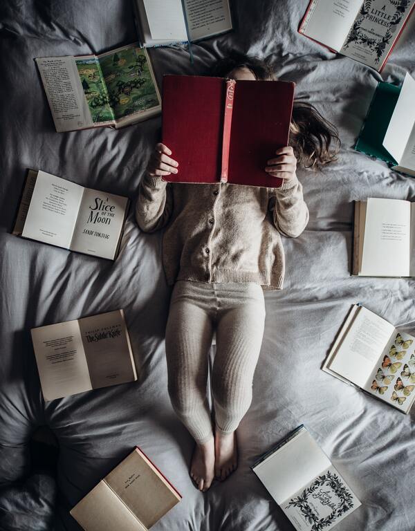 Uma mulher deitada com um livro aberto e vários outros livros abertos ao redor dela. A foto foi tirada de cima. 