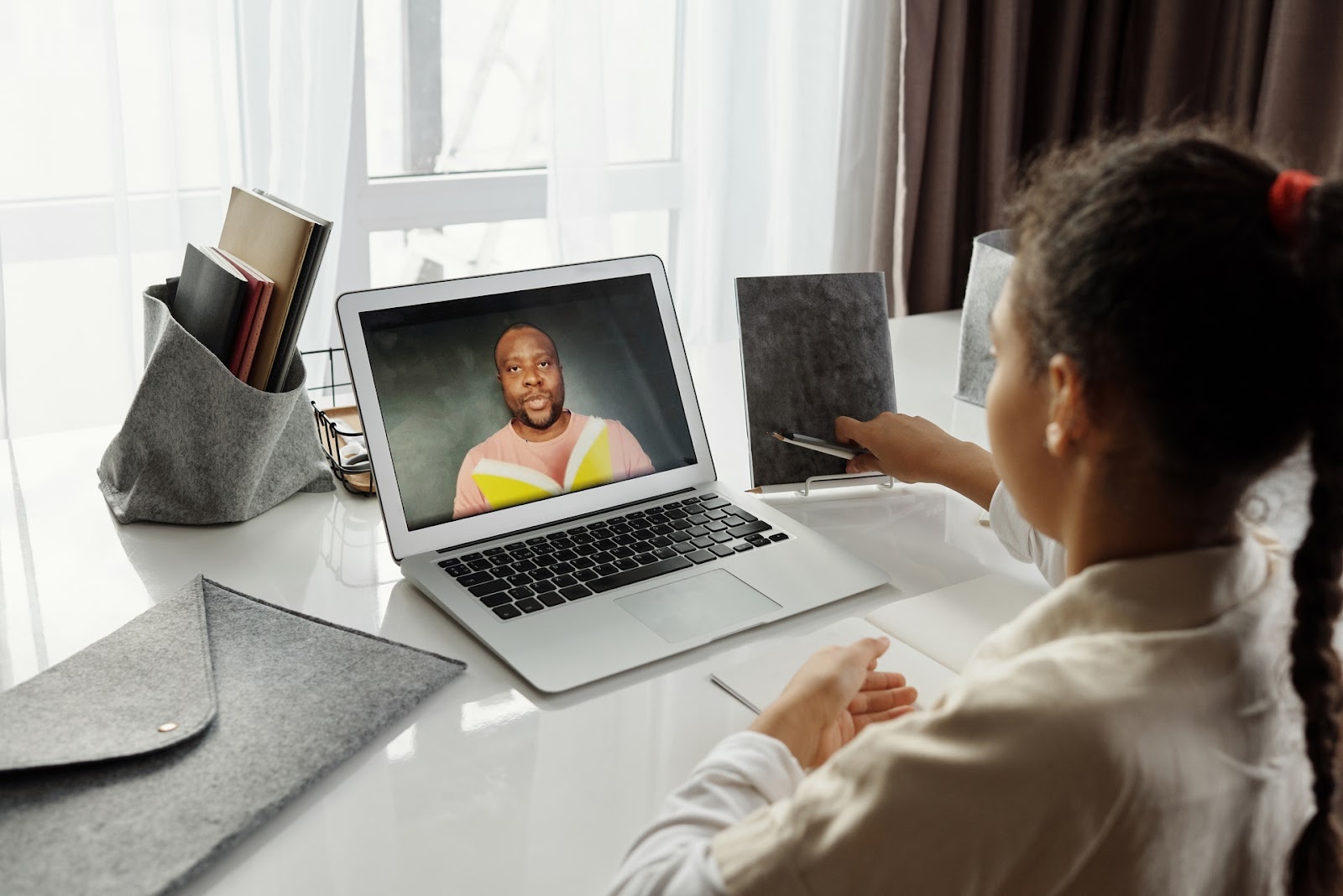 A student sits at a white desk while having a video conference with a man on her computer screen. She has books and a notebook on her desk.