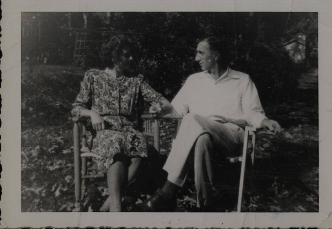 Black and white photo of Opal Weimer, seated in a rocking chair wearing a floral dress and beaded necklace, and  Fred Tice, seated in a rocking chair wearing a button up shirt and pants. The couple are looking at each other.