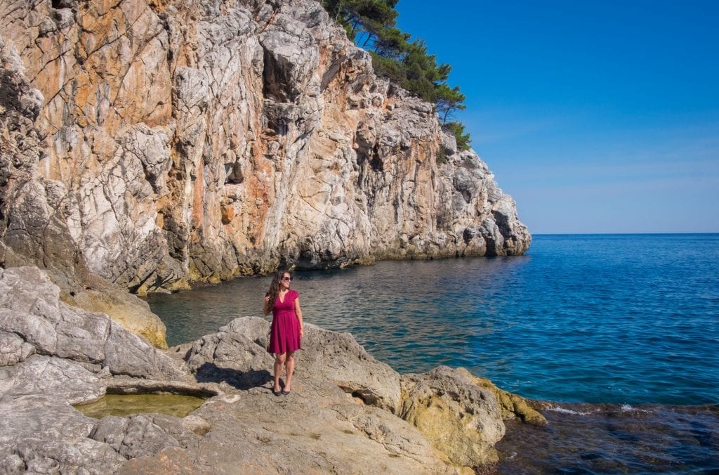 Kate standing on the edge of the rocky coastline in Dubrovnik, wearing a red dress and sunglasses, looking out to sea.