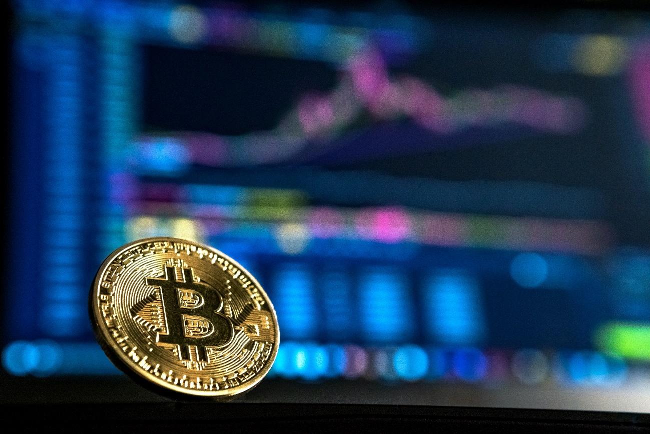Bitcoin representada por uma moeda física. Atrás aparecem gráficos que dão ideia da volatilidade das criptomoedas.