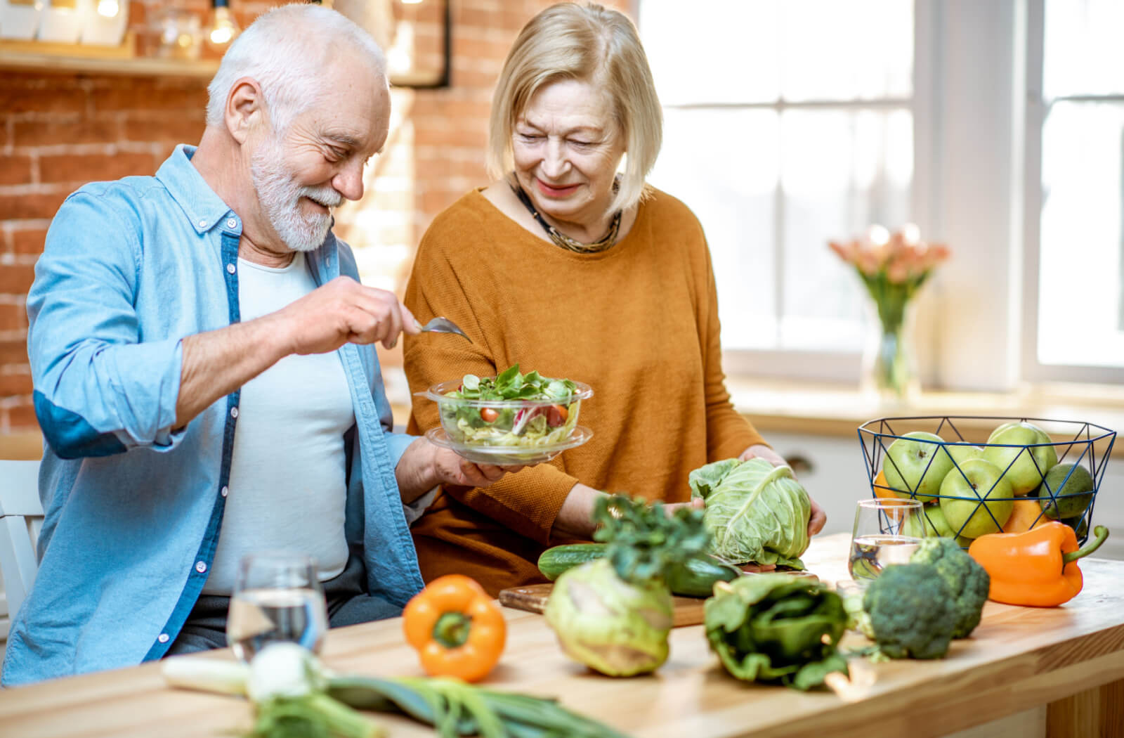 A senior man and a senior woman preparing a bowl of salad in the kitchen.