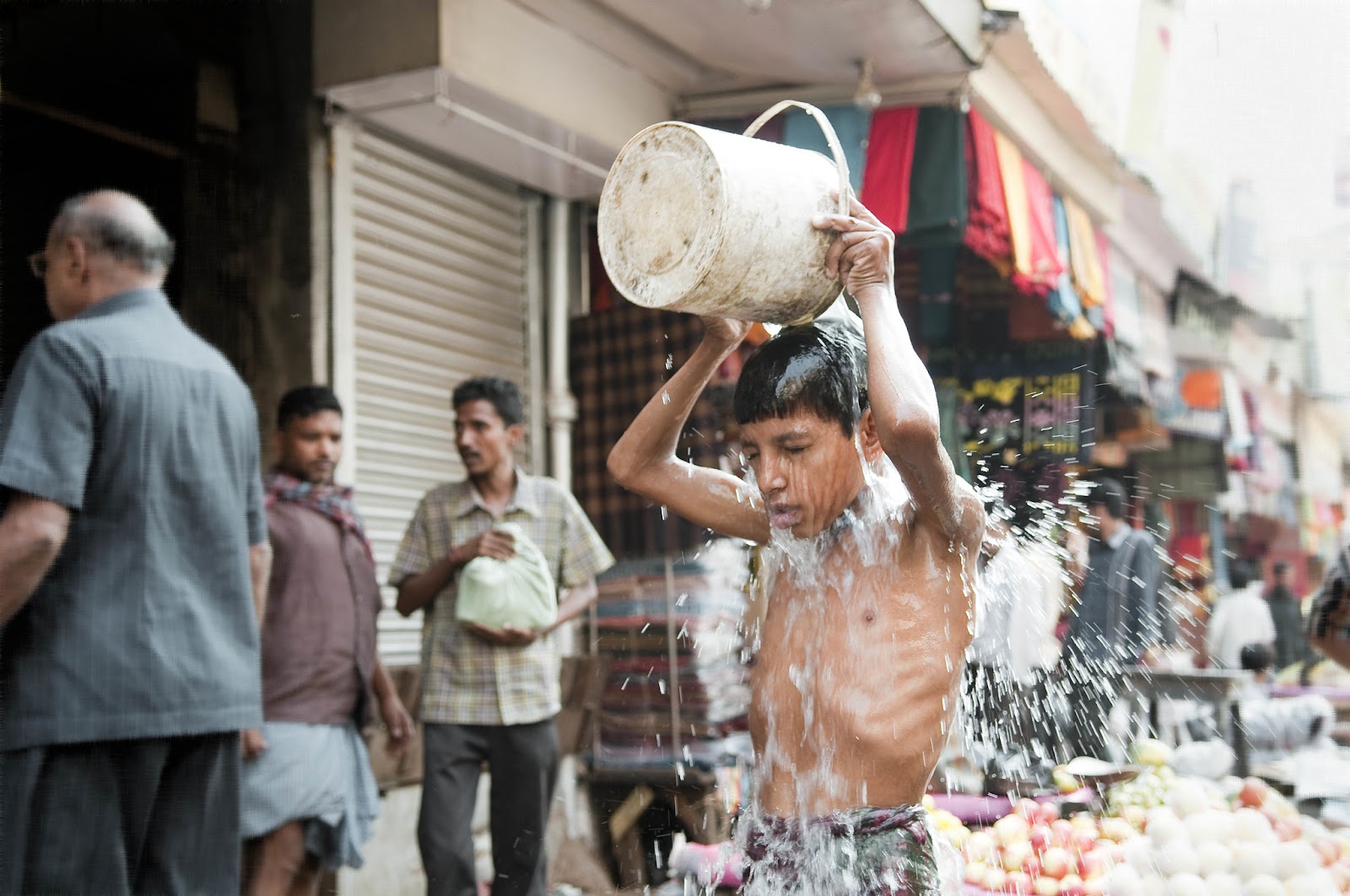 Zdjęcie przedstawia chłopca na ulicy, który wylewa na siebie wiadro wody.
