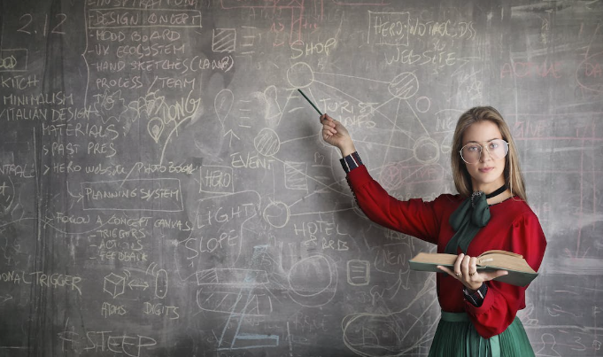 A teacher is teaching on a blackboard. She has a book in her hand.


