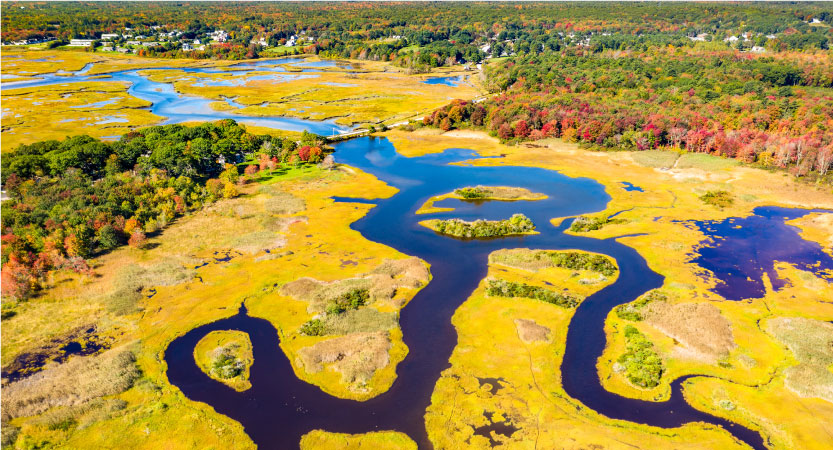 Aerial view of Little River estuary in Wells, Maine, in the summer. In the distance are residential homes and lush woodlands.