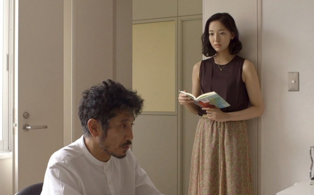 A woman in a brown shirt holds a book while a man in a white shirt sits
