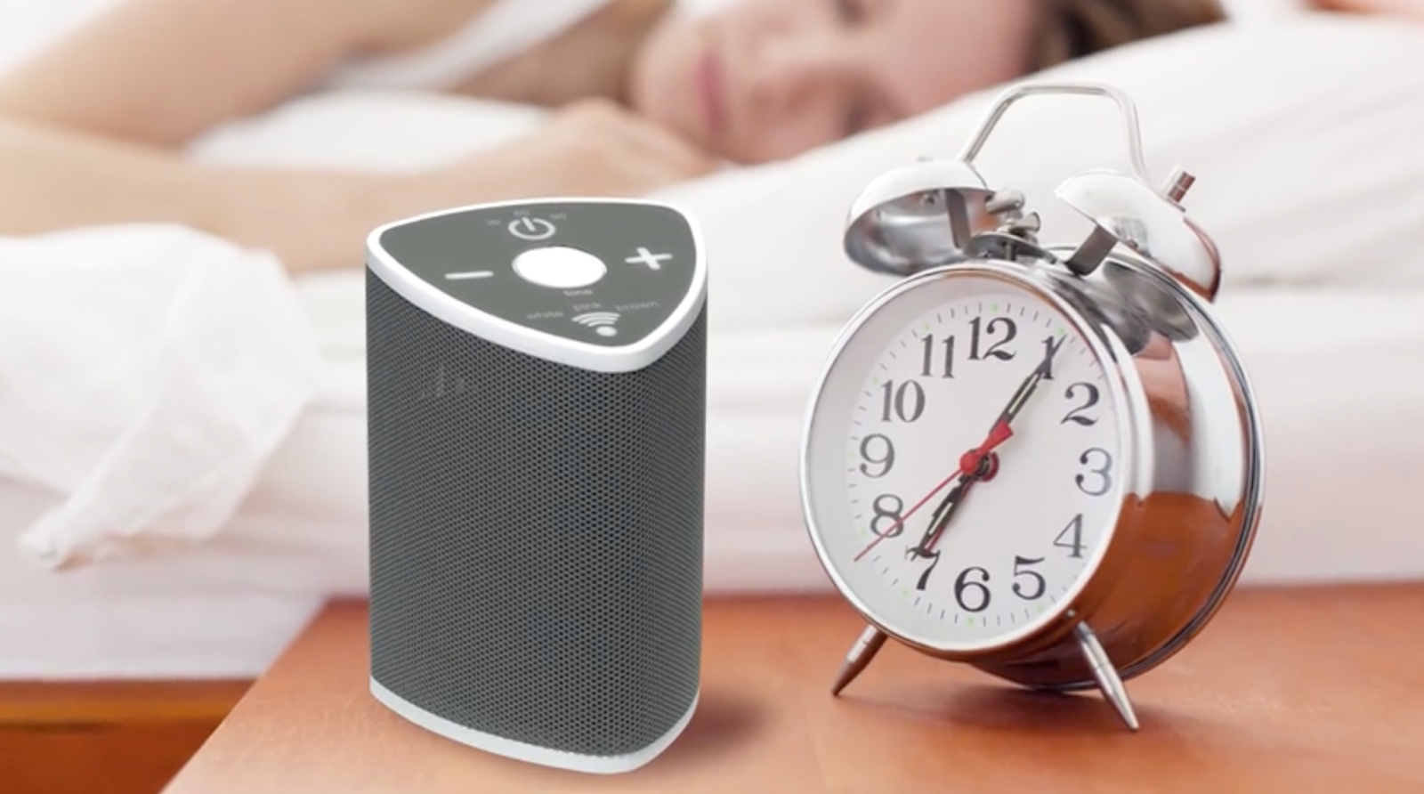A Sonorest Sleep Tones Sound Machine  for tinnitus on a bedside table next to an alarm clock. A woman is asleep in the background.