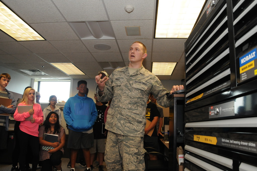 An Air Force officer in fatigues stands in the front of the room addressing a small group of high school students, some of whom are holding books and notebooks.