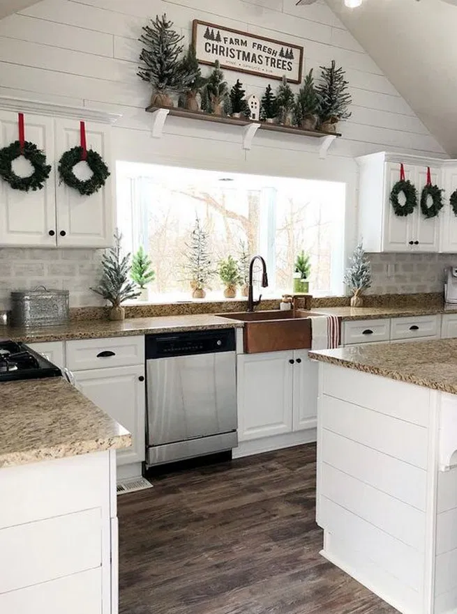 farmhouse christmas kitchen decor featuring christmas wall sign, mini cabinet wreaths and mini christmas trees on the countertops