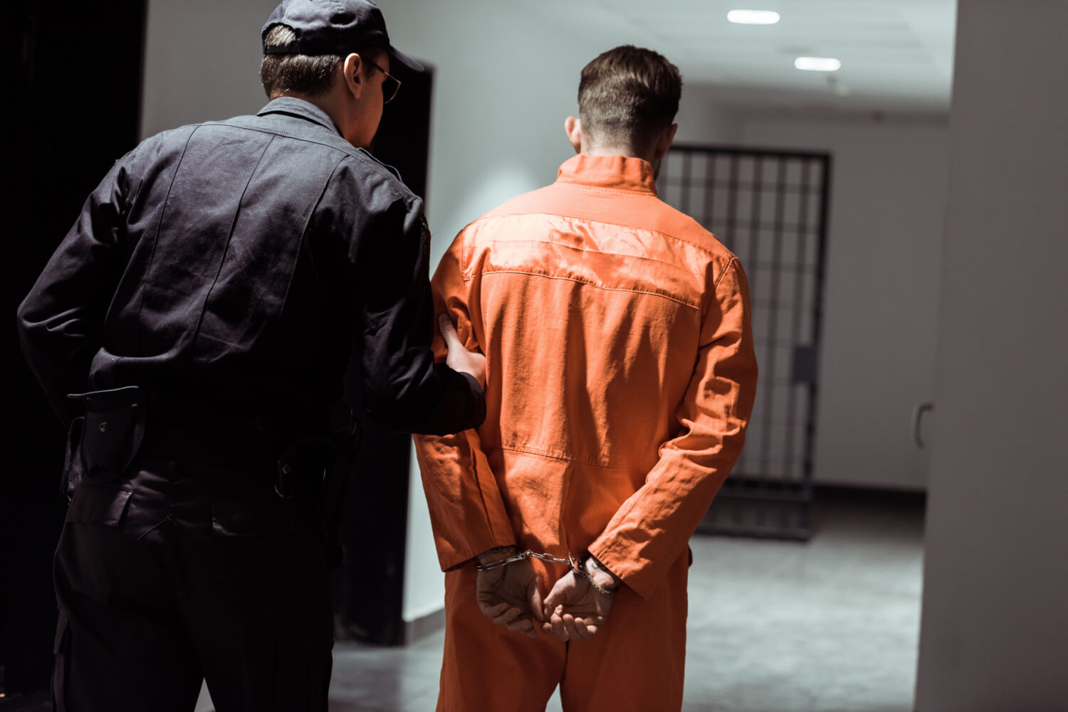 vista traseira de um agente penitenciário conduzindo um prisioneiro algemado em um corredor  
