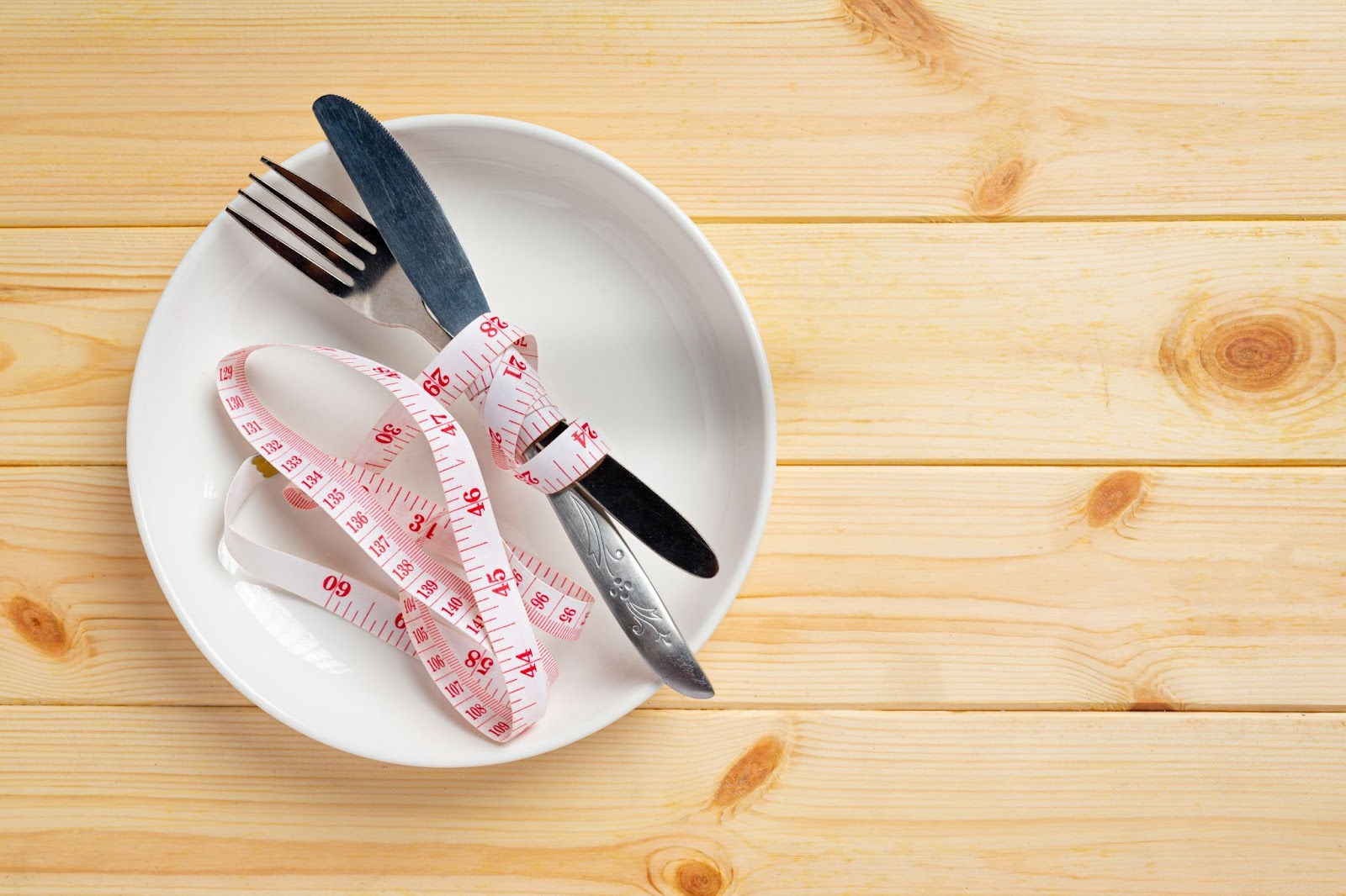 Visão de cima da mesa de madeira com prato branco redondo apenas com um garfo e uma faca cinzas amarrados por uma fita métrica branca com numeração vermelha