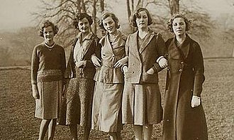 Nearly full-length group portrait of five well-dressed women standing in a field. Their ages range from roughly 20 to 30; their hair is cut short of the shoulders in elegant 1930s or 1940s styles; four of the five wear skirts down just below the knee, and one a longer coat. Two wear pearls.
