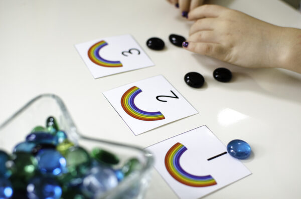 A bin filled with multi-colored flat marbles in the bottom left corner.  In the middle of the table are three cards: the first numbered 1, the second numbered 2, and the third numbered 3 at the bottom of each card. There is a rainbow at the top of each card. Under card one is one marble, under card two is two marbles, and under card three, a child is placing three flat marbles.