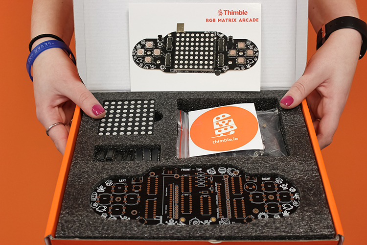 An Thimble kit box is held by two hands, open, and displaying the components and instruction cards for the RGB Matrix Arcade STEM kit.