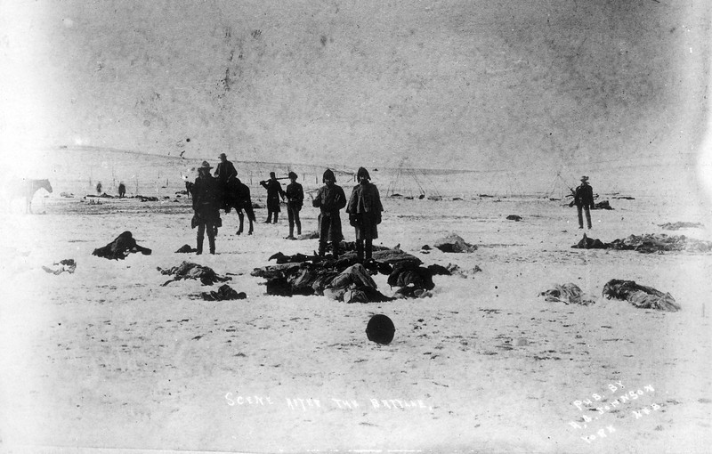 Description of  "Scene after the battle." Jan. 1891. View southwest from center of council circle after the fight at Wounded Knee Creek, Pine Ridge Reservation, South Dakota, shows men holding moccasins and other souvenirs among the frozen bodies of Native American Lakota Sioux on the snow covered ground. (Denver Public Library; Western History Collection)