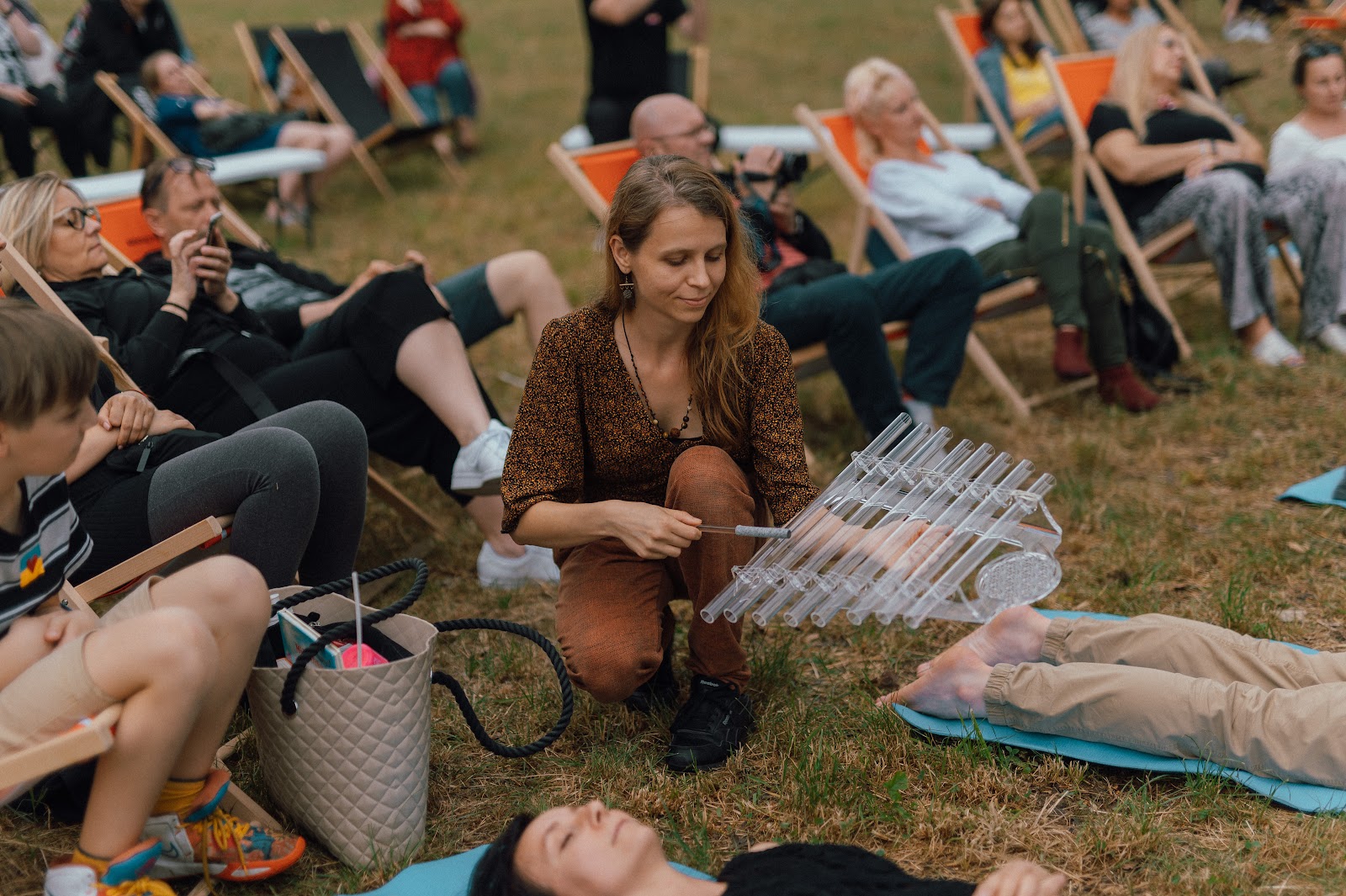 Młoda kobieta trzymająca w ręce szklany instrument przypominający cymbałki kuca pośród ludzi siedzących na leżakach oraz leżących na matach na trawie. Trwa koncert relaksacyjny.