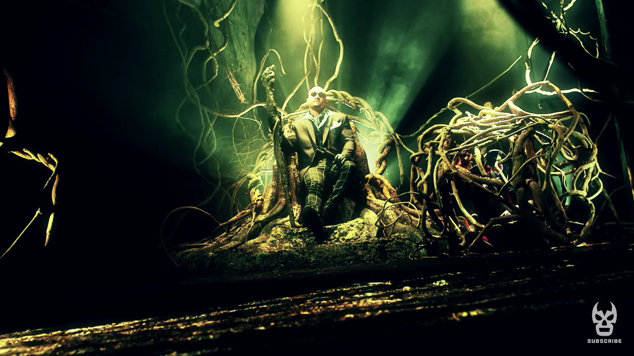 Screen cap from Lucha Underground. Killer Kross, a white man with a clean-shaven head, is wearing sunglasses and a suit and clutching a knobby stick. He's sitting on a throne made of twisted tree branches in an eerie green forest and to his right is a ball of twisted up tree branches housing a prisoner, another luchador. 