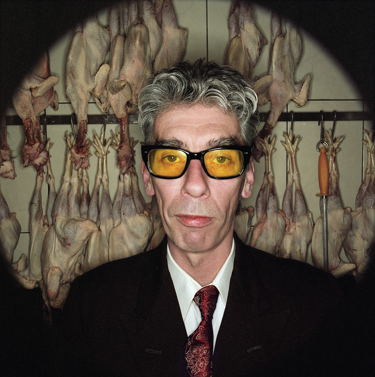 A quirky image by photographer Matt Carr with the personality of the subject shining through. The subject, an older gentleman with salt and pepper hair and a serious expression, wears a fairly formal black suit and red tie. However, his formal image is offset by the bright yellow tinted lenses in his glasses and the chicken carcasses hanging in the background.
