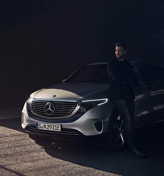 In a dark street, the light hits a silver Mercedes Benz, while a guy stands next to it looking cool and pensive. 
Photo by Klubhouse Collective