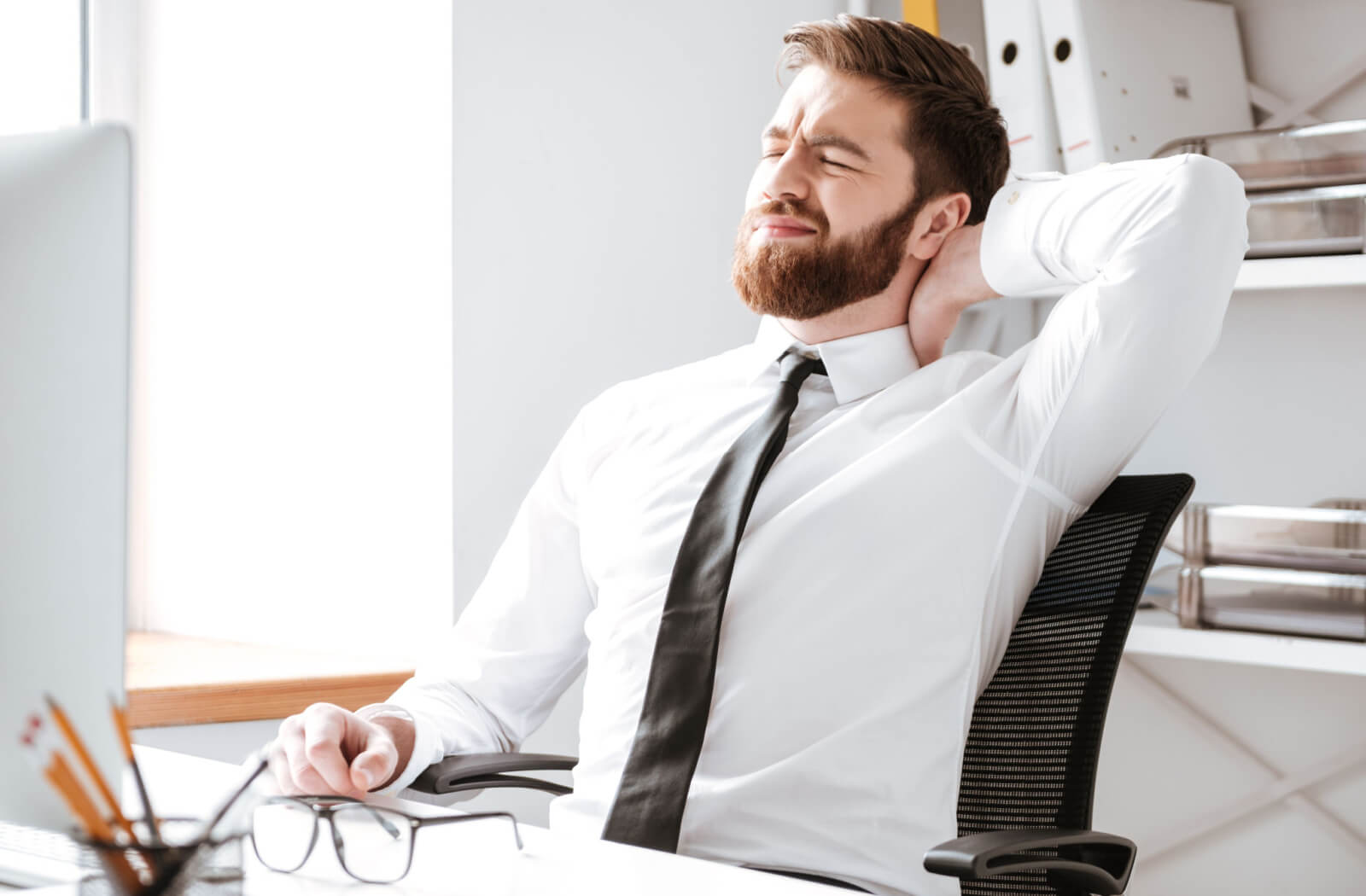 A young man in a white collared long sleeve and a black necktie is sitting on his black office chair with his left hand touching his neck and making some stretching to relieve his neck and Eye Strain