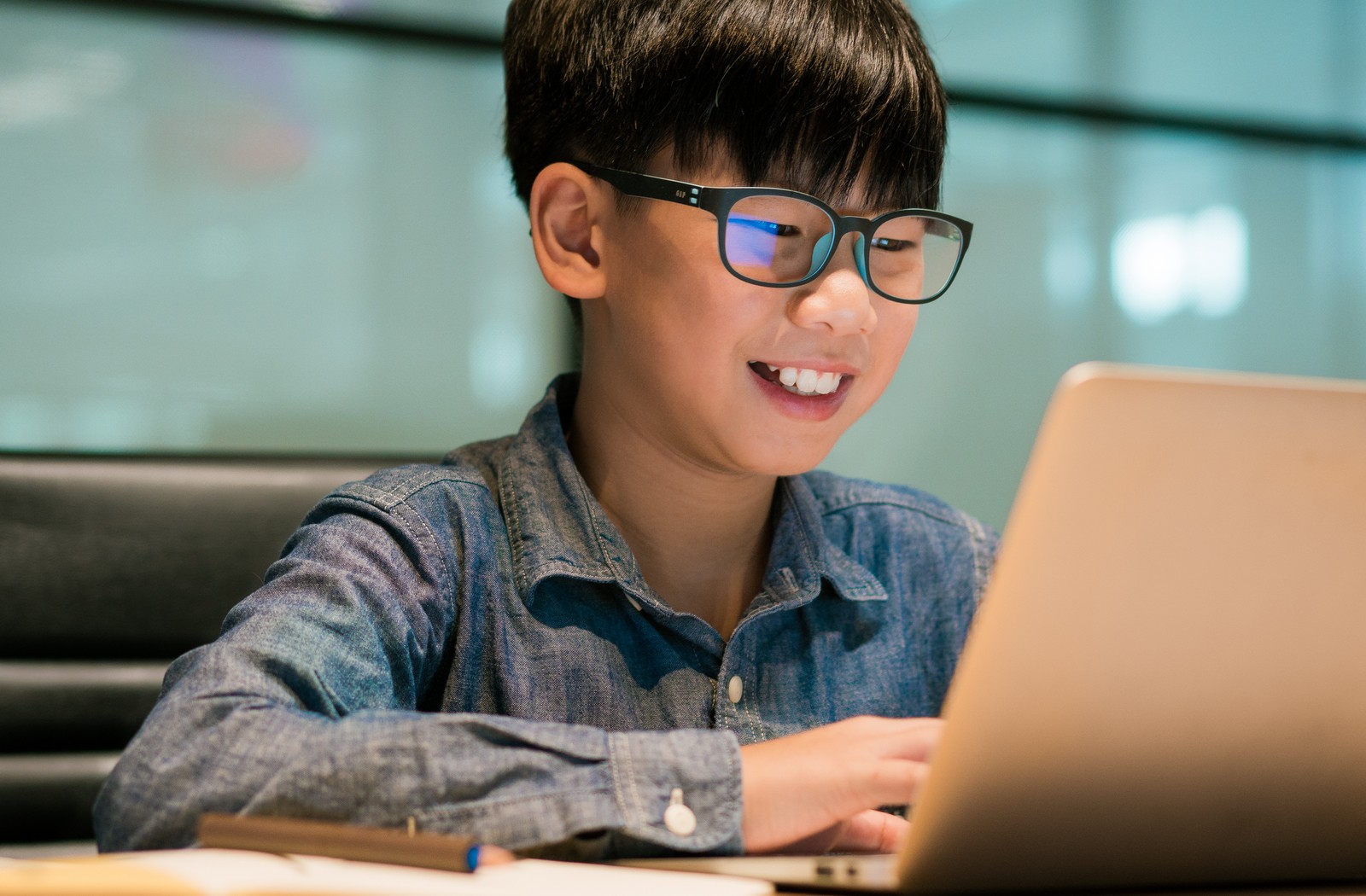 Pre-teen asian boy wearing dark rimmed glasses and denim shirt smiling while staring at his laptop computer on his desk