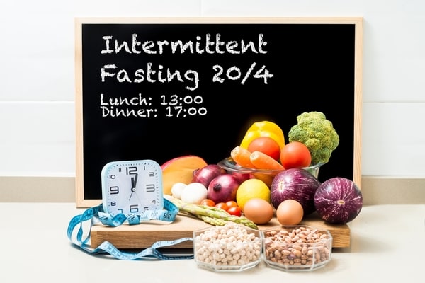 A small square-shaped analog clock, blue tape measure, and healthy foods on a wooden board with a black board that says "Intermittent Fasting 20/4"