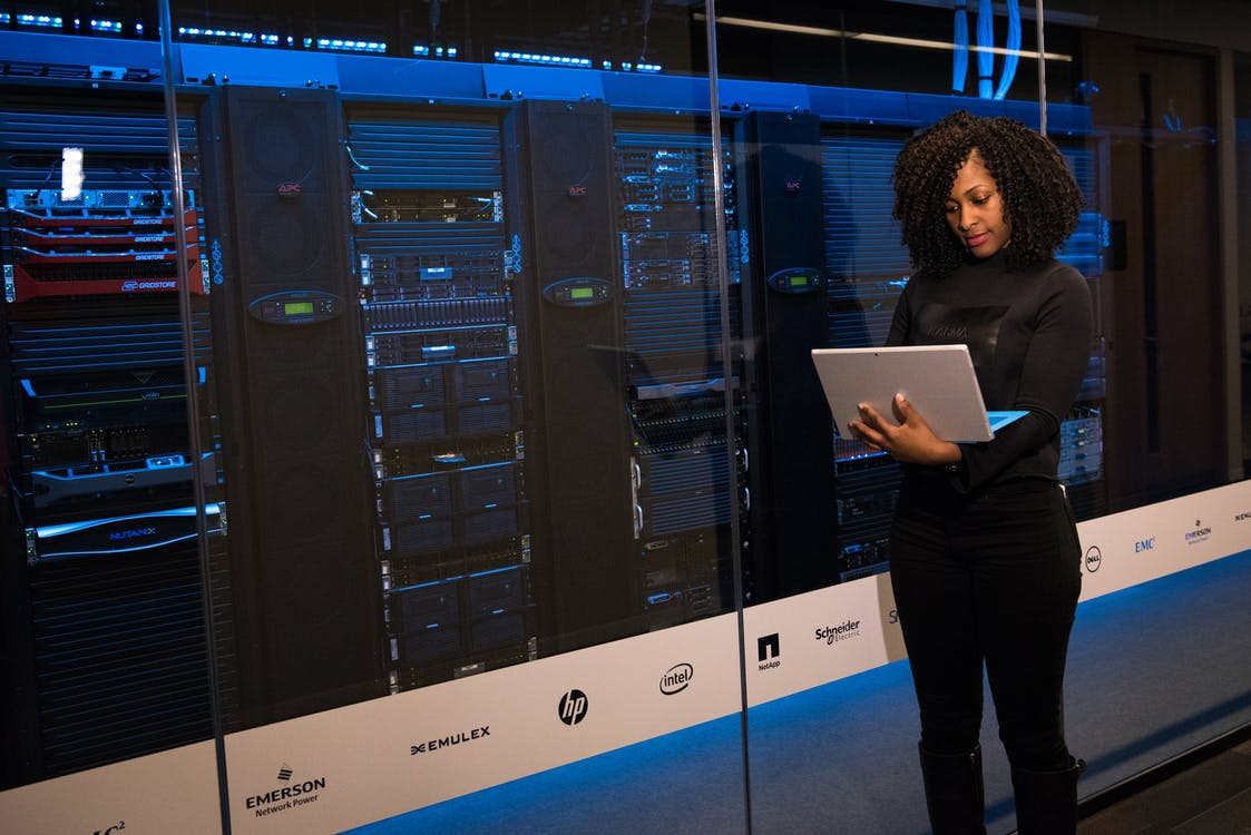 A woman holding a laptop and standing next to a data server