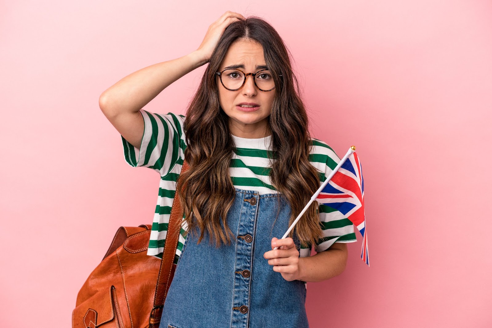 a Caucasian brown hair woman in glasses holding the UK flag and scratching her head on pink background