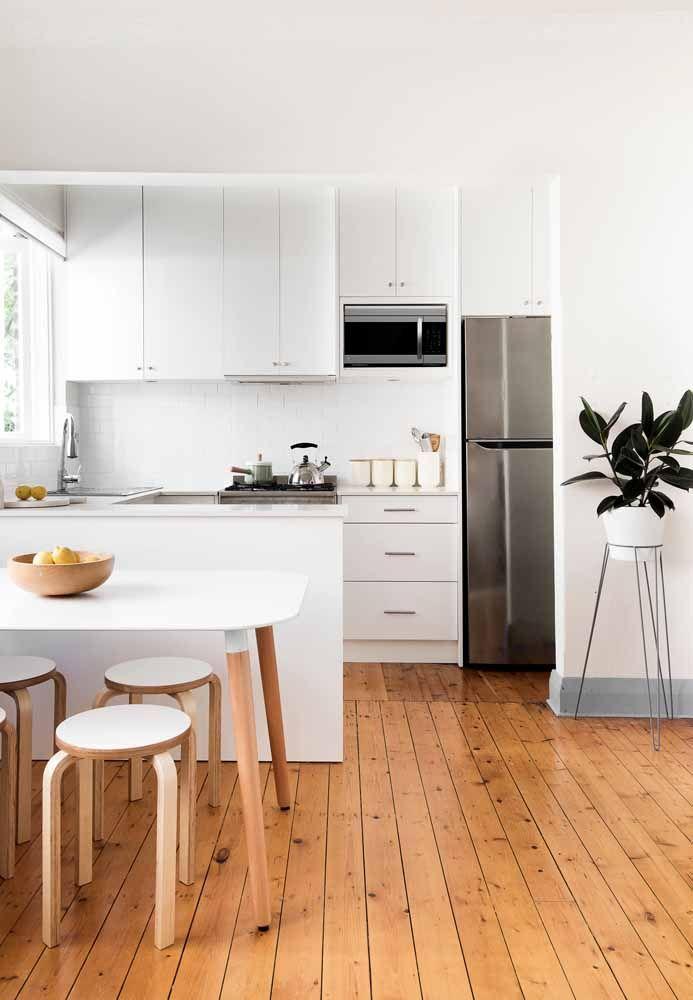 Cozinha com armários brancos, eletrodomésticos brancos, bancada e mesa branca, bancos brancos com estrutura de madeira, piso de madeira e vaso de planta branco