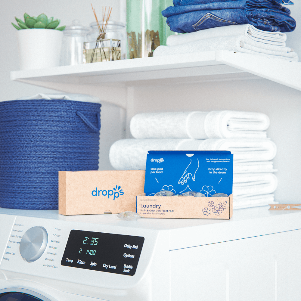 Feeling Fresh and Cleaning Up the Laundry Industry with Dropps–Two boxes of Dropps’ laundry detergent pods sitting on top of a white washing machine. There are clean, folded towels, a blue wicker laundry basket, and a shelf with various decor items above the washing machine.  