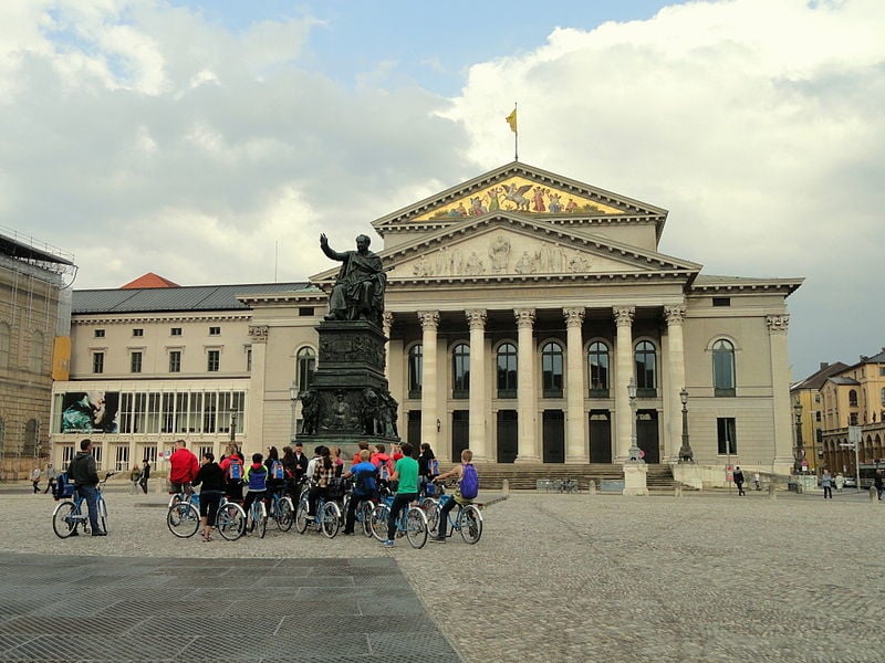 Tour guide with tourists on bicycles standing under a statue in Max-Joseph-Platz.