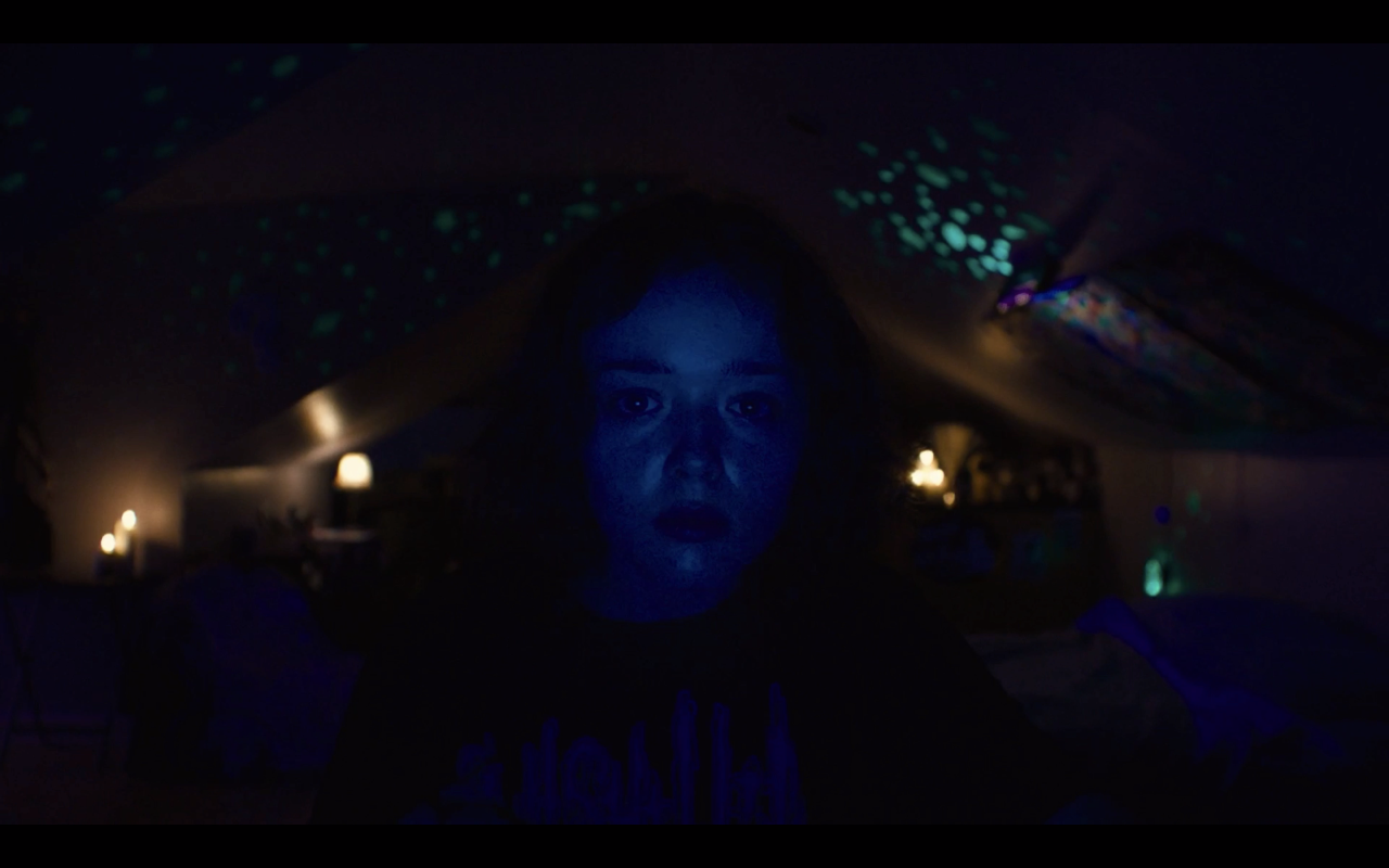 A still from We're All Going to the World's Fair. Casey sits in front of her laptop camera in a dark attic bedroom. Colored lights and lamps are lit behind her.