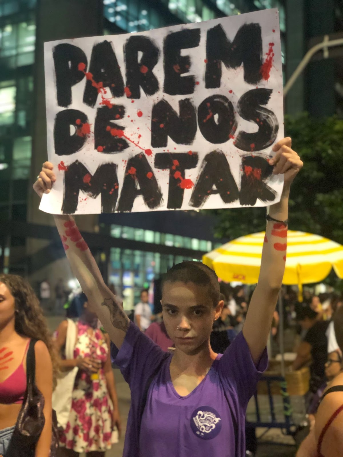 Foto colorida de um protesto com uma mulher levantando um cartaz dizendo: “Pare de nos matar”
