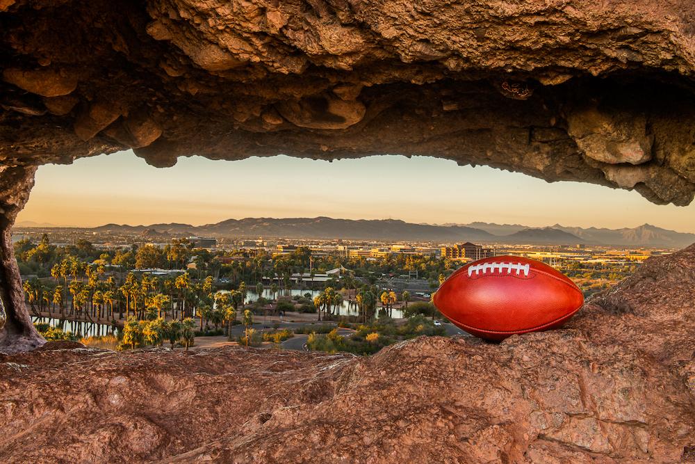 Image of a brown leather football in the foreground positioned in peekhole rock formation overlooking trees and reflective water, with dusky mountains on the horizon by outdoor/adventure photographer Aaron Ingrao. 