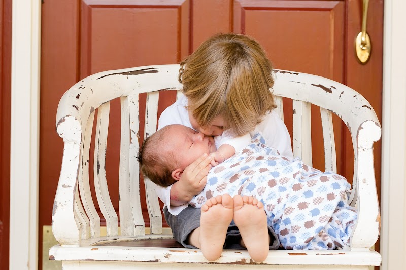 A toddler sits on a white chair outside against a red door, the toddler is holding and kissing a swaddled baby