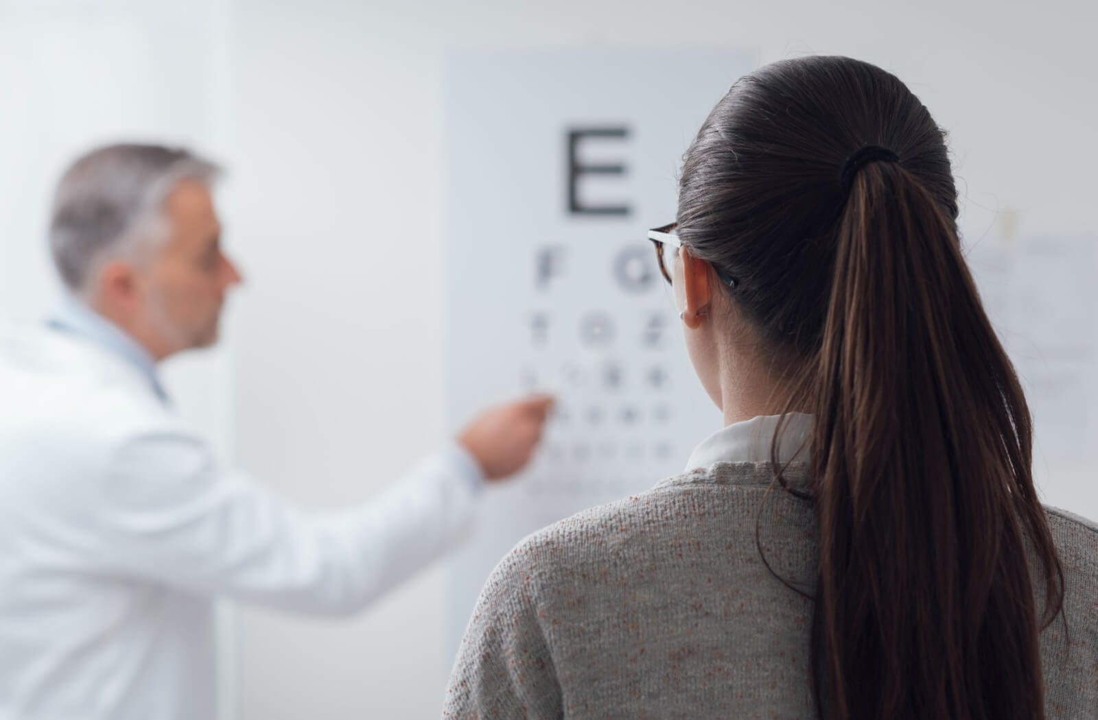 A lady in a brown-colored sweat shirt is facing far from the wall while the optometrist is pointing something into the Snellen chart.