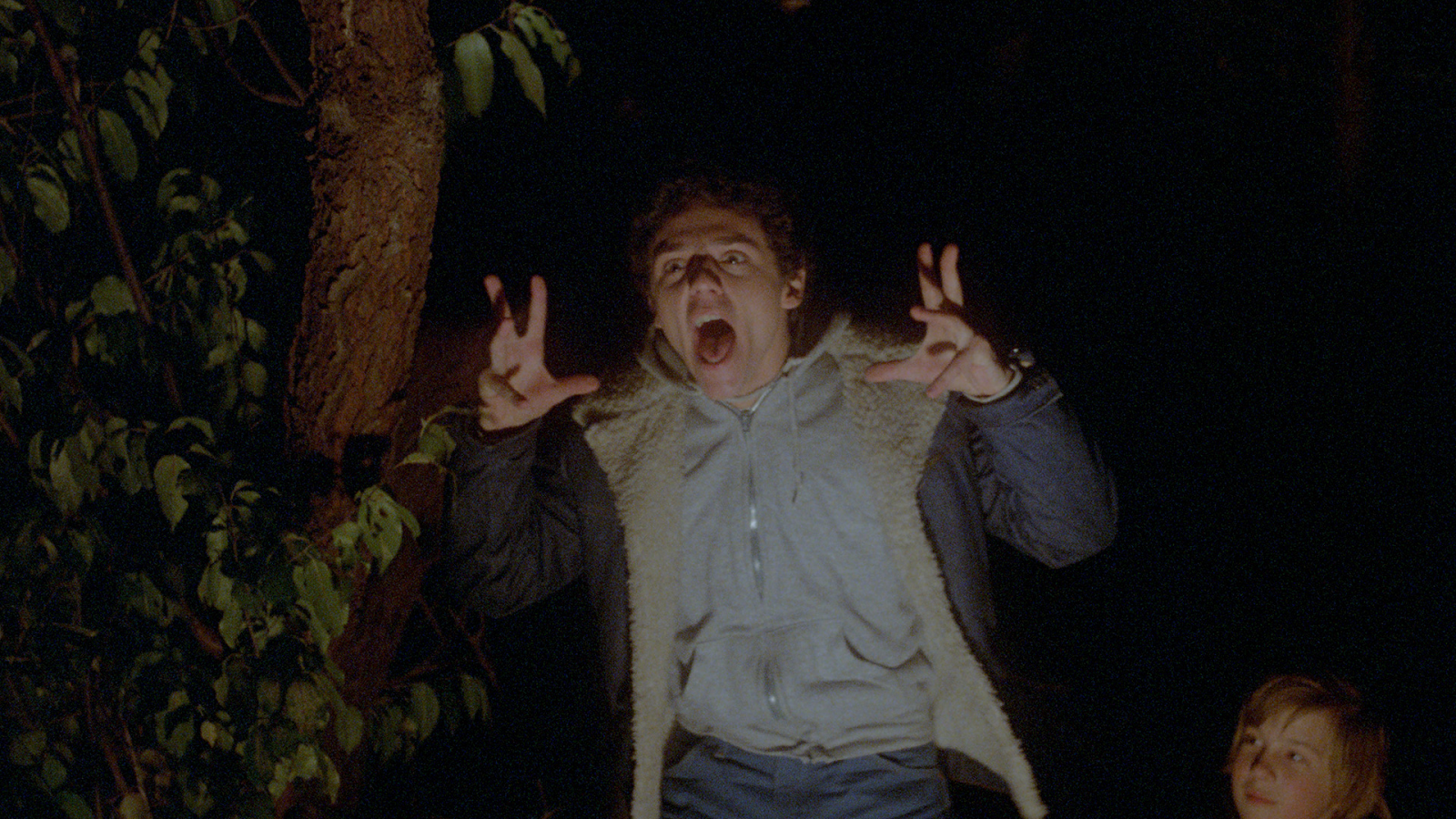 Still from Madman (1981). A teenage boy acting scary in the woods, making a face and raising his hands in clawing motions. A small boy sits beside him, watching. 